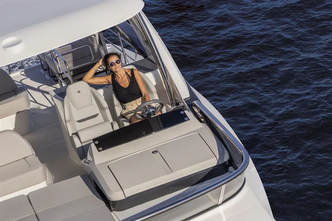 A woman relaxes at the helm of a sleek yacht, wearing sunglasses, with the sea in the background and modern seating nearby.
